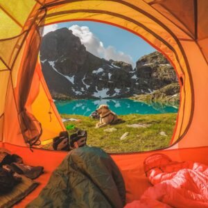 View from inside an orange tent of a picturesque mountain scene. A lake with turquoise water and surrounding rocky mountains can be seen. A dog is sitting just outside the tent, with some camping gear, sleeping bags, and a hat visible inside the tent.