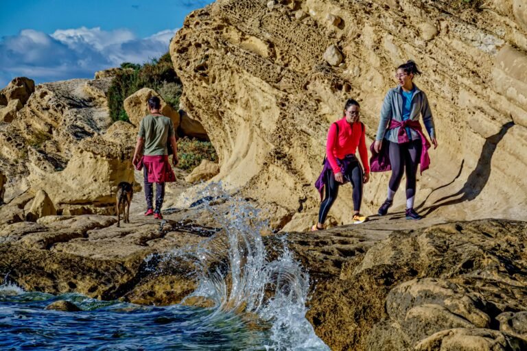 Three people and a dog are standing on rocky terrain near crashing waves. The group appears to be exploring or hiking, with one person looking out at the water, and two others near a large rocky formation. The sky is partly cloudy and the sea is vibrant.