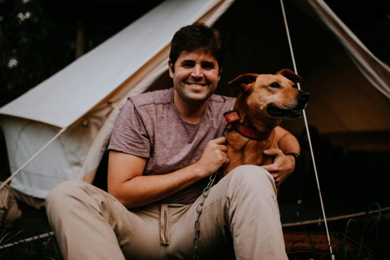 A man wearing a purple shirt and beige pants sits in front of a tent, smiling and holding a brown dog with a red collar. The tent is set up in an outdoor, grassy area, suggesting a camping setting.