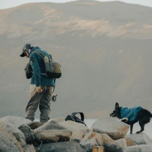 A person wearing fishing gear and a cap stands on rocky terrain near a body of water, holding a fishing rod with a line cast. A dog in a blue vest walks on the rocks beside them. Mountainous landscape is visible in the background under an overcast sky.