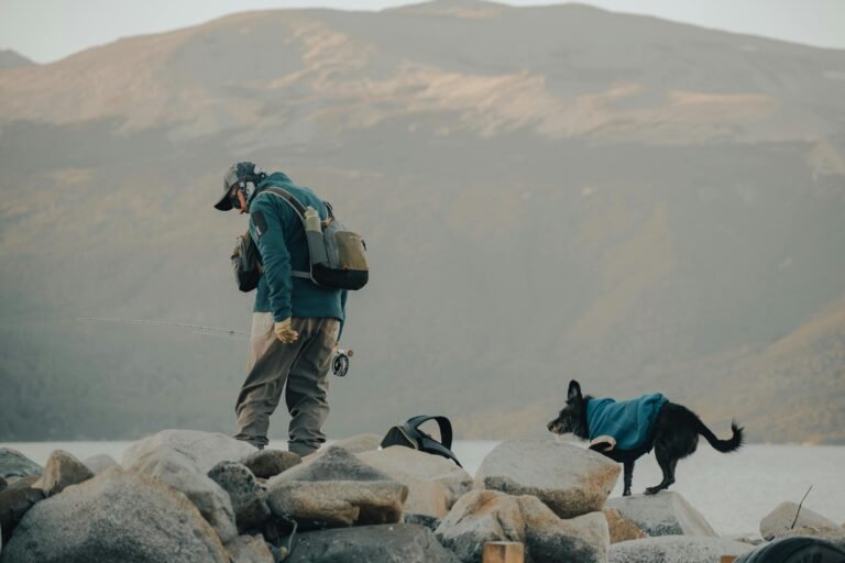 A person wearing fishing gear and a cap stands on rocky terrain near a body of water, holding a fishing rod with a line cast. A dog in a blue vest walks on the rocks beside them. Mountainous landscape is visible in the background under an overcast sky.