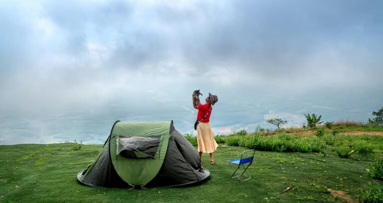 A person wearing a red shirt and beige skirt lifts a small dog into the air while standing next to a green camping tent and a blue folding chair. They are on a grassy hillside with a misty mountain landscape in the background.